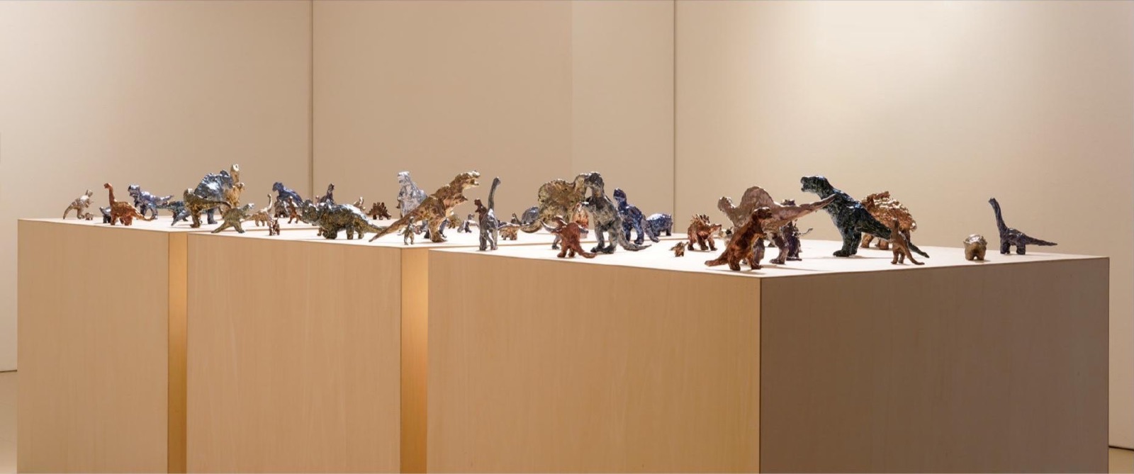 絵具で覆われた多数の恐竜フィギュアを白い台上に並べた展示風景